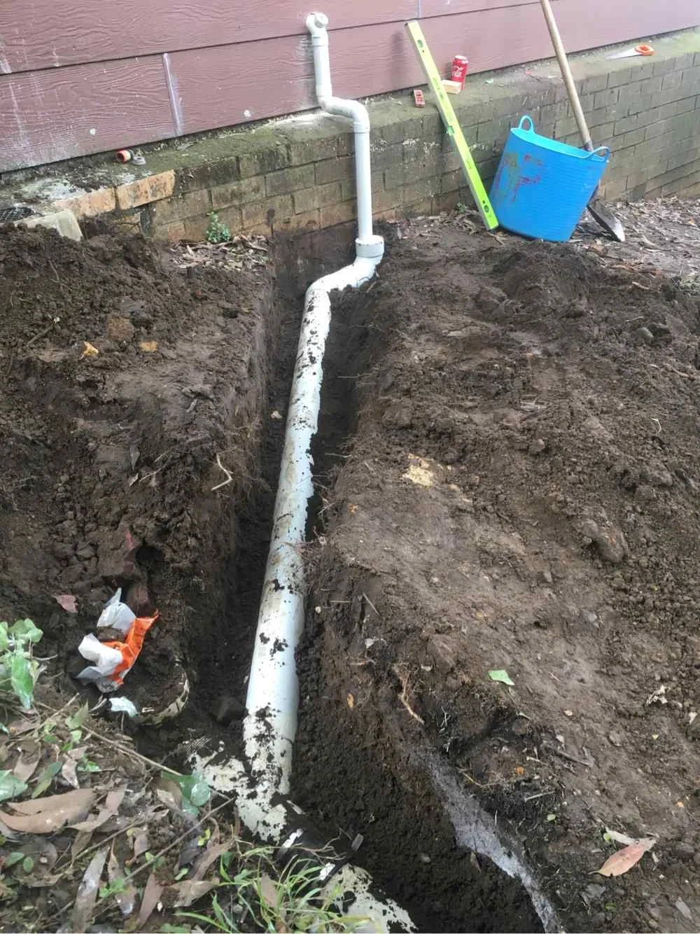 New drainage pipe laid in a narrow trench beside a brick wall at a residential property