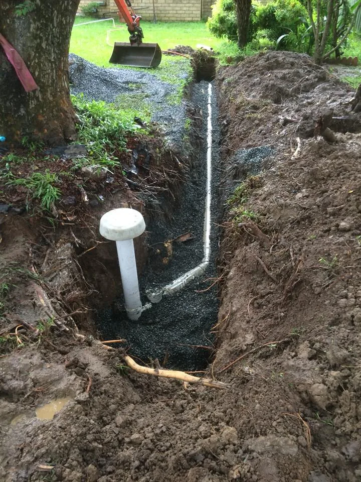Gravel-lined drainage trench with a new pipe and inspection cap installed beside a tree root at a residential property