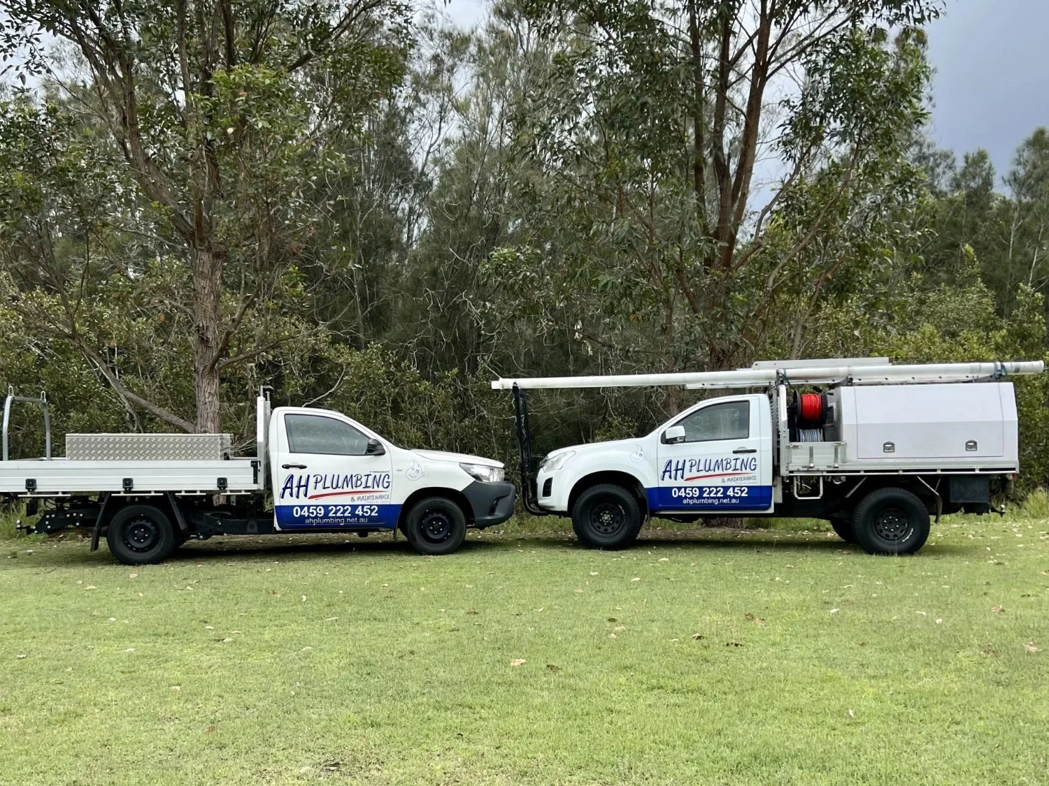 Two AH Plumbing & Maintenance branded utes parked on grass in front of bushland