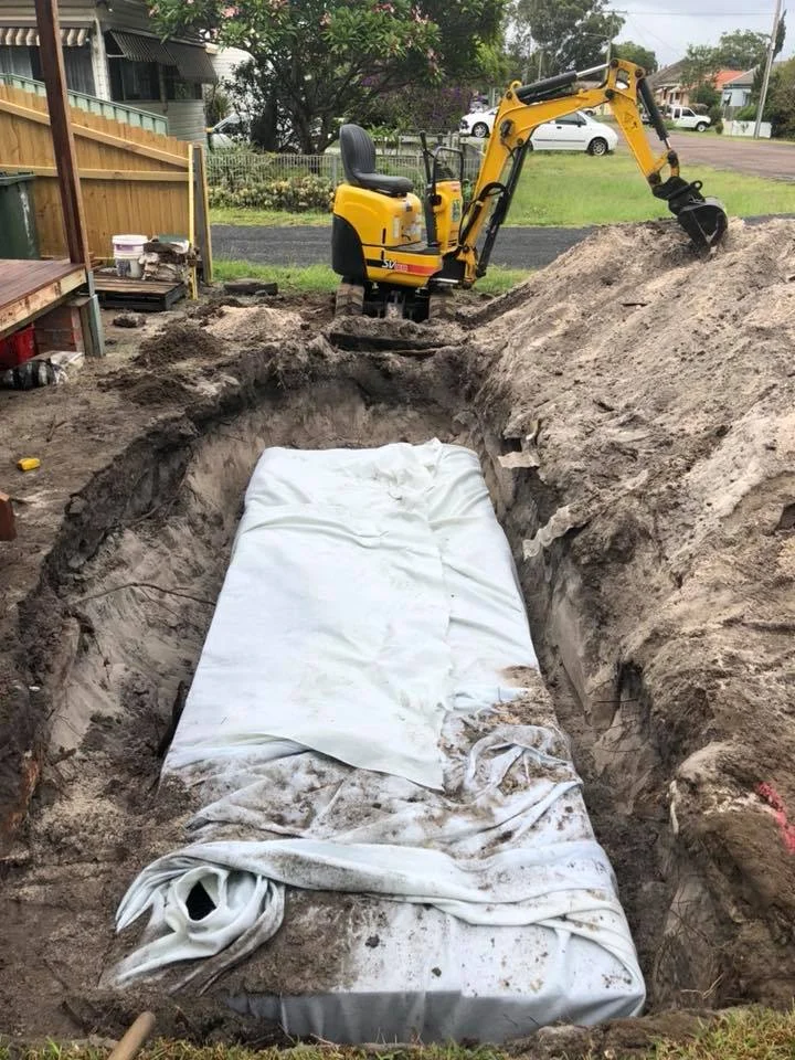 Mini excavator beside a deep excavation trench wrapped in geotextile fabric at a residential property