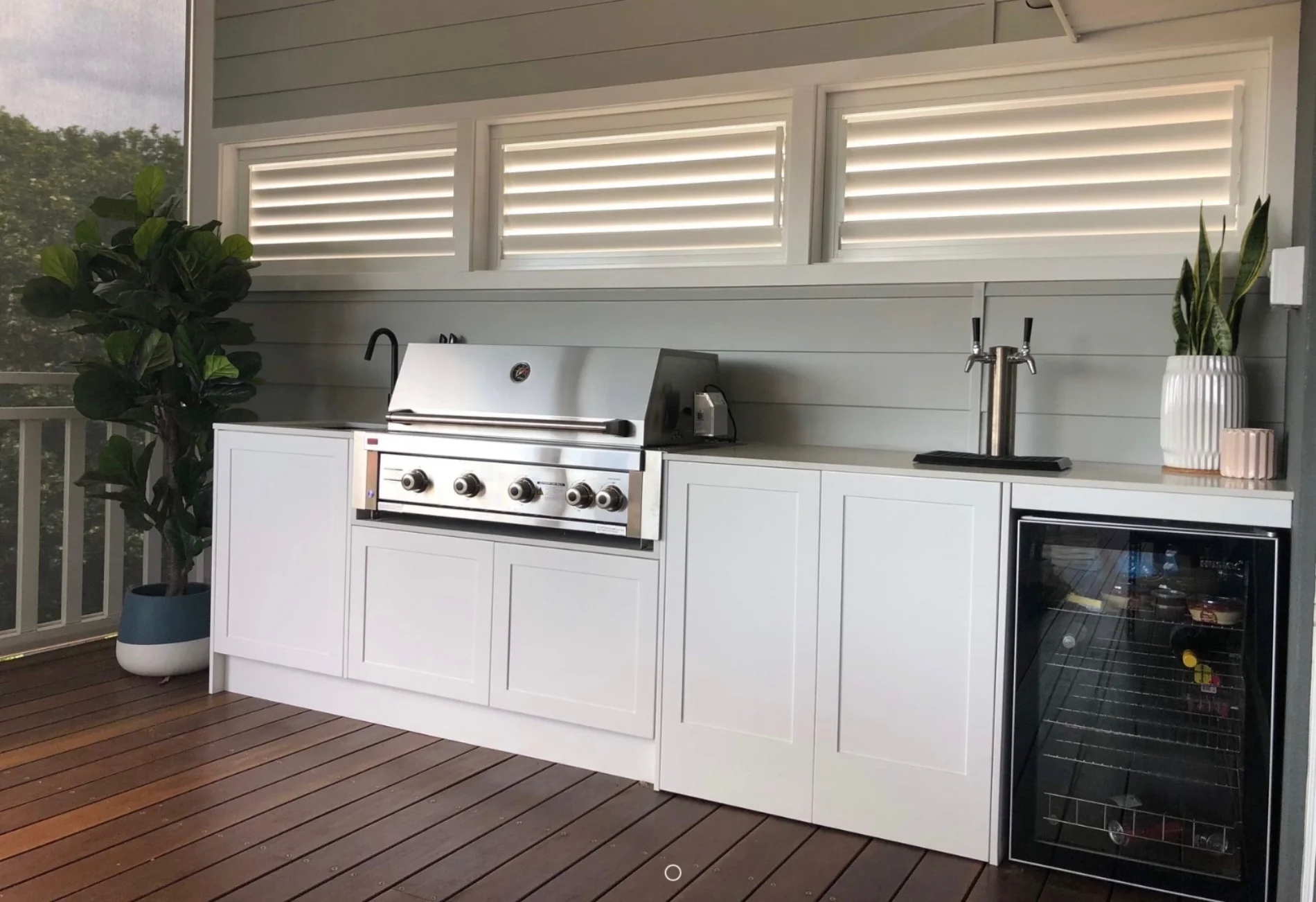 Outdoor kitchen with built-in gas BBQ, white cabinetry and bar fridge installed on a timber deck by AH Plumbing & Maintenance