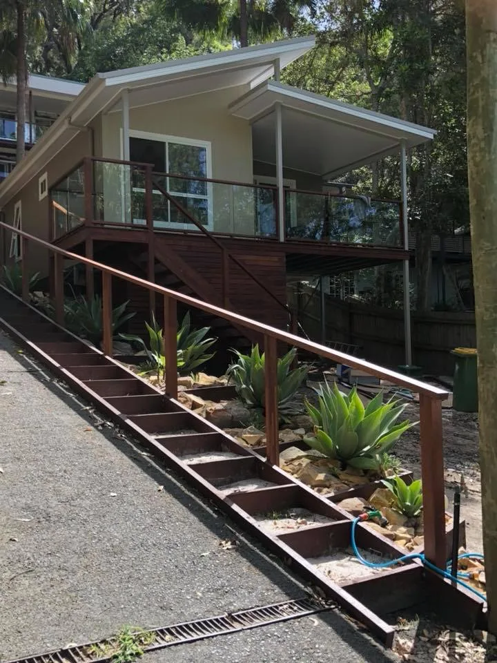 Elevated residential home with a timber deck, glass balustrade and pipework running along the driveway